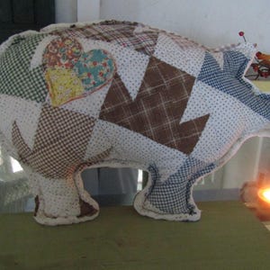 May include: A stuffed pig-shaped pillow constructed from various fabric patches in brown, white, blue, and green. The patchwork features plaid, gingham, and polka dot patterns, with a prominent floral heart appliqué on the side of the body.
