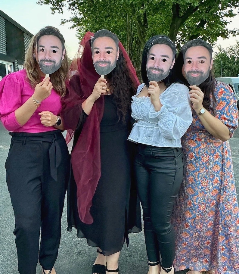 May include: Four people are wearing paper masks of the same man's face. The people are wearing different outfits, including a pink top and black trousers, a maroon scarf and black dress, a white top and black trousers, and a floral dress.