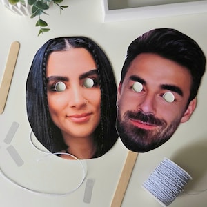 May include: Two paper face masks with eye holes. The masks are of a man and a woman. The woman's mask has long black hair and the man's mask has short brown hair.