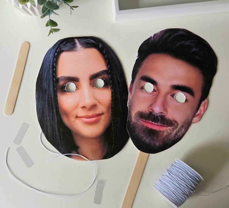 May include: Two paper face masks with eye holes. The masks are of a man and a woman. The woman's mask has long black hair and the man's mask has short brown hair.