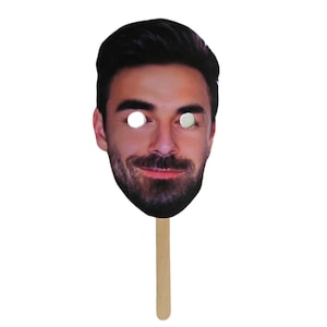 May include: A cardboard face mask with a photo of a man with a beard and a stick attached to the bottom.