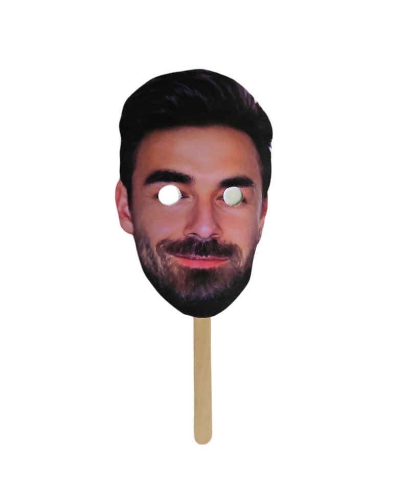 May include: A cardboard face mask with a photo of a man with a beard and a stick attached to the bottom.