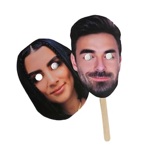 May include: Two cardboard face masks on sticks. The masks are of a man and a woman. The man has brown hair and a beard. The woman has long dark hair. Both masks have white circles for eyes.