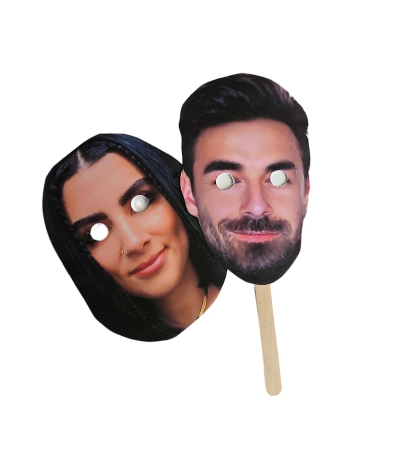 May include: Two cardboard face masks on sticks. The masks are of a man and a woman. The man has brown hair and a beard. The woman has long dark hair. Both masks have white circles for eyes.