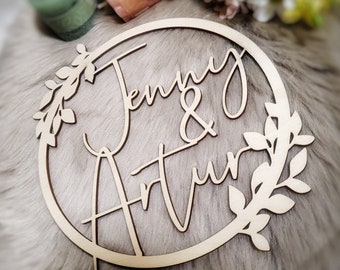 May include: A wooden circle with the names "Jenny & Arthur" written in cursive script. The names are surrounded by a leafy wreath.
