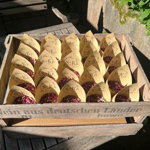 May include: A wooden crate labeled Wein aus deutschen Landern containing multiple tan paper cones filled with dried pink and red flower petals. Each cone features a printed wreath design with the initials S and K and a date in the center.
