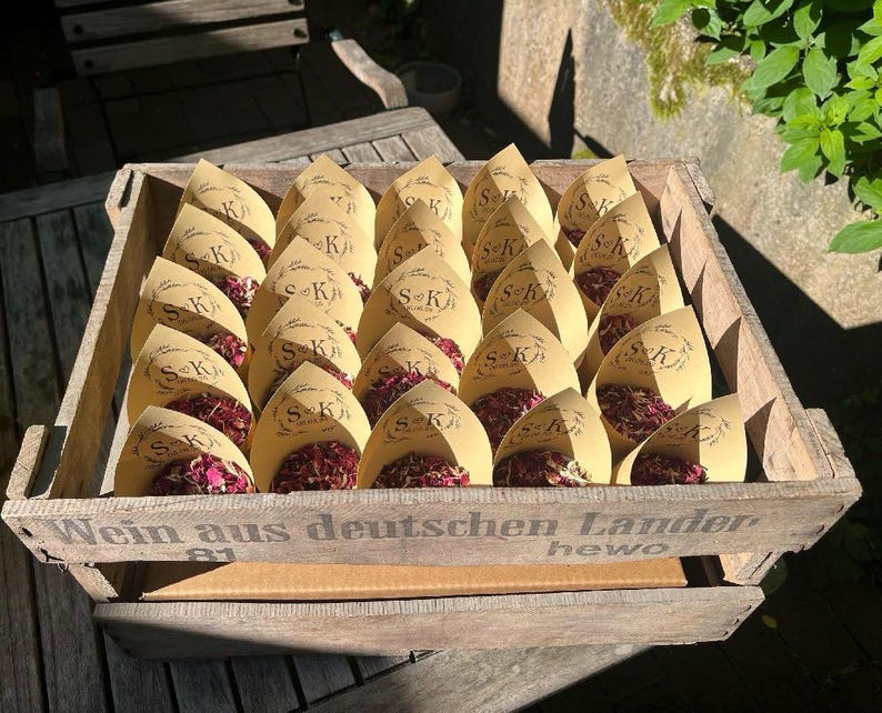 May include: A wooden crate labeled Wein aus deutschen Landern containing multiple tan paper cones filled with dried pink and red flower petals. Each cone features a printed wreath design with the initials S and K and a date in the center.