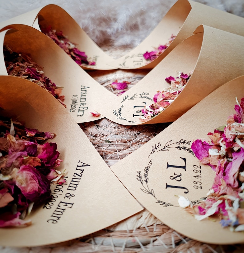 May include: Several light brown paper cones filled with dried pink, red, and white flower petals. Each cone features black printed text including names like Arzum & Emre or initials J&L, along with specific dates such as 10/06/2022 or 23.4.22.