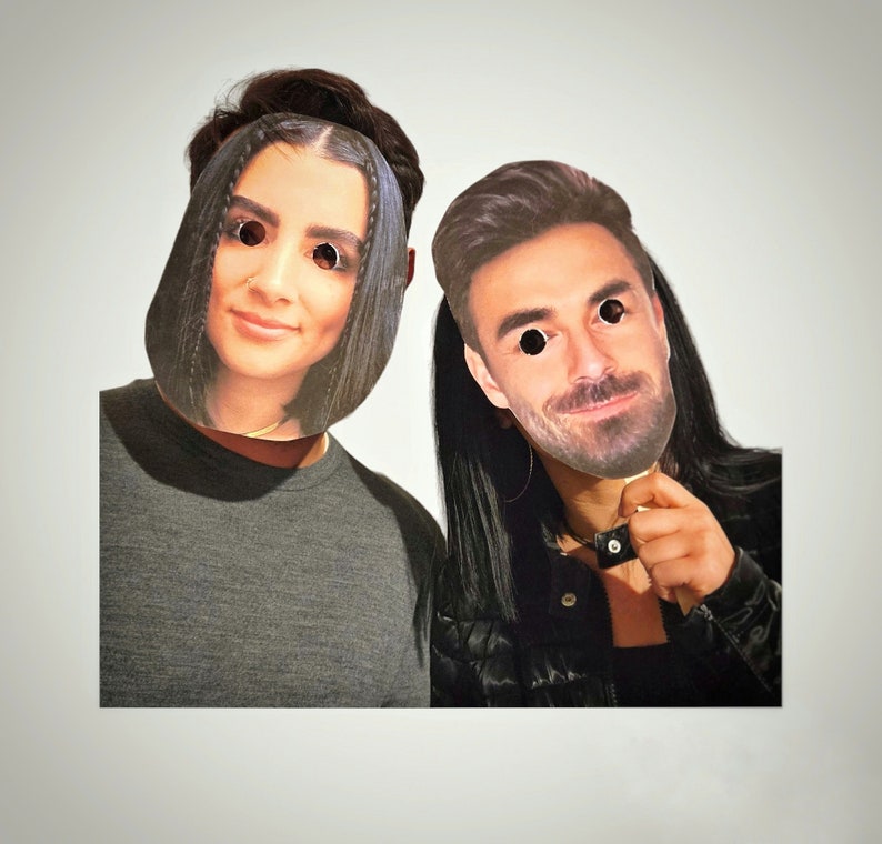 May include: Two people are wearing paper masks of faces. The person on the left is wearing a grey shirt and the person on the right is wearing a black jacket.