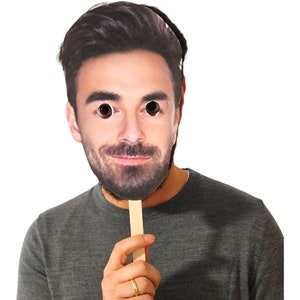 May include: A cardboard face mask with a printed image of a man's face. The mask is attached to a wooden stick.