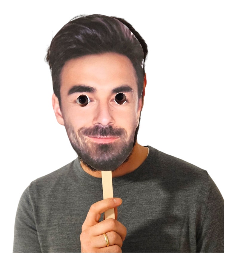 May include: A cardboard face mask with a printed image of a man's face. The mask is attached to a wooden stick.