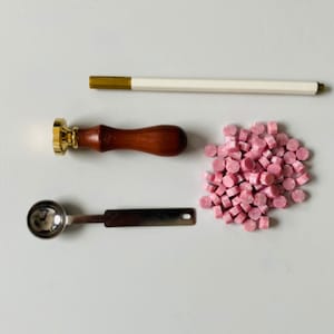 K&ouml;nnte beinhalten: Ein Set mit Wachssiegelzubeh&ouml;r, bestehend aus einem Wachssiegel mit Holzgriff, einem kleinen L&ouml;ffel aus Metall, einem Wachsstab und einem Haufen rosa Wachsperlen.
