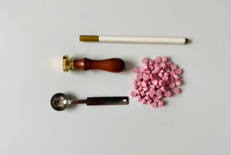 K&ouml;nnte beinhalten: Ein Set mit Wachssiegelzubeh&ouml;r, bestehend aus einem Wachssiegel mit Holzgriff, einem kleinen L&ouml;ffel aus Metall, einem Wachsstab und einem Haufen rosa Wachsperlen.