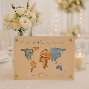 Op de afbeelding: Een houten wereldkaart met uitgesneden continenten, beschilderd in blauw, oranje en bruin. De tekst "Die schönste Reise beginnt zu zweit" staat bovenaan, met de namen "Alina & Stefan" en de datum "18.05.2026" onderaan. Goudkleurige studs bevestigen de kaart.