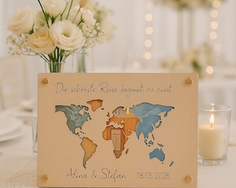 May include: A wooden world map with cut-out continents, painted in blue, orange, and brown, is displayed. The text "Die schönste Reise beginnt zu zweit" is at the top, with names "Alina & Stefan" and the date "18.05.2026" at the bottom. Gold-colored studs secure the map.