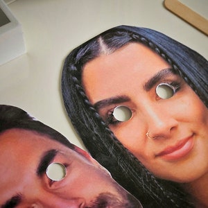 May include: Two paper masks with eye holes cut out. The masks are of a woman with long dark hair and a man with short dark hair.