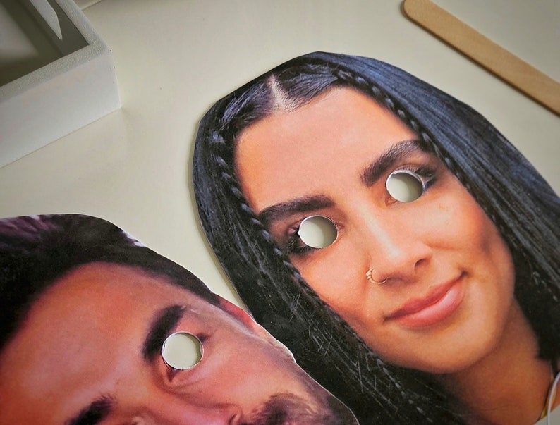 May include: Two paper masks with eye holes cut out. The masks are of a woman with long dark hair and a man with short dark hair.