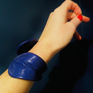 May include: A close-up of a person's arm wearing a dark blue leather bracelet. The bracelet is made of multiple layers of leather and has a slightly wrinkled texture.