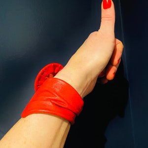 May include: A red leather bracelet with a large, round, folded design. The bracelet is worn on a person's wrist, and their thumb is pointing up.