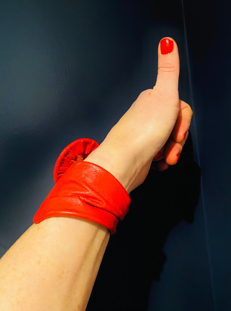 May include: A red leather bracelet with a large, round, folded design. The bracelet is worn on a person's wrist, and their thumb is pointing up.