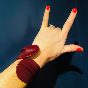 May include: A maroon leather cuff bracelet with a unique design. The bracelet is made of two pieces of leather that are joined together at the wrist.