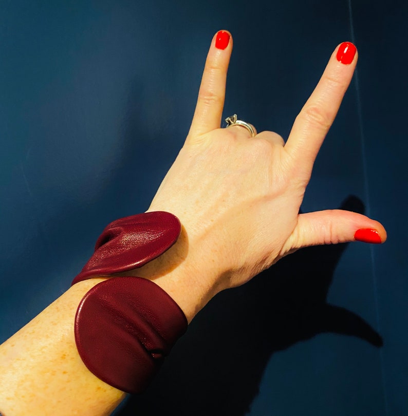 May include: A maroon leather cuff bracelet with a unique design. The bracelet is made of two pieces of leather that are joined together at the wrist.