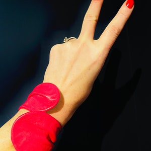 May include: A pink leather bracelet with two large, rounded, overlapping pieces. The bracelet is worn on a person's wrist.