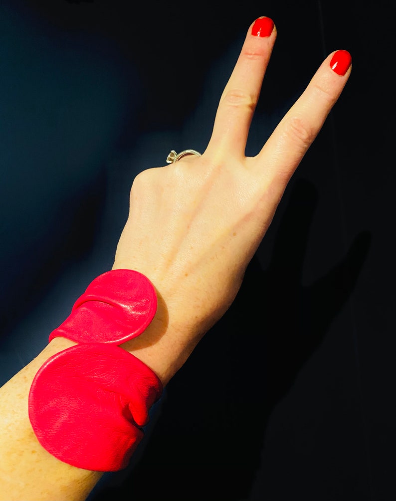 May include: A pink leather bracelet with two large, rounded, overlapping pieces. The bracelet is worn on a person's wrist.