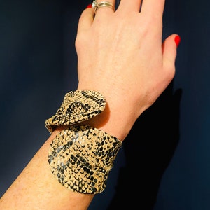 May include: A beige and black snakeskin patterned cuff bracelet. The bracelet is made of leather and has a unique, twisted design.