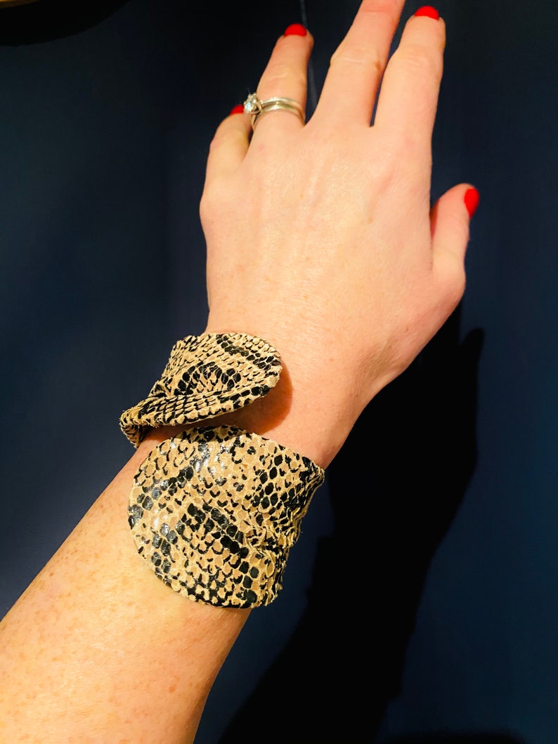 May include: A beige and black snakeskin patterned cuff bracelet. The bracelet is made of leather and has a unique, twisted design.