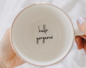 May include: White ceramic mug with a gold rim and the text "hello gorgeous" printed on the inside.