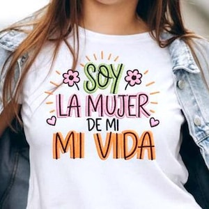 Puede incluir: Camiseta blanca con la frase en español "SOY LA MUJER DE MI VIDA" en letras coloridas. El diseño incluye detalles florales y corazones. Una prenda de vestir casual.
