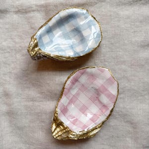 Peut inclure: Deux coquilles d'huîtres décoratives avec des bords peints en or. L'une présente un motif vichy bleu et blanc, l'autre un motif vichy rose et blanc. Les coquilles sont posées sur un fond neutre.