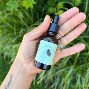 May include: A small amber glass dropper bottle with a black cap and a light green label. The label features a black line drawing of a person and the text "THE PRIMAL HERB" and "BRAIN TONIC" printed in a dark green font on the front surface.