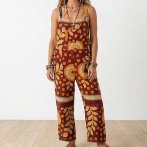 May include: A burgundy jumpsuit with a floral and dot pattern. The jumpsuit features adjustable straps, a bib pocket, and wide legs. The design includes yellow and orange leaf and sunburst motifs.