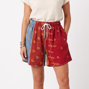 May include: A pair of shorts featuring a patchwork design with panels of red, blue, and yellow fabric. The shorts have a white drawstring waist, an elastic waistband, and a repeating pattern of small yellow leaf and geometric motifs throughout.