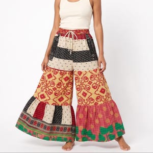 May include: Wide-leg patchwork pants in a variety of colors and patterns. The pants feature a drawstring waist and a tiered, ruffled hem. The patchwork includes red, black, cream, and yellow fabrics with geometric and floral designs.