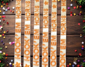 May include: Beaded ribbons with the text "HOOK 'EM HORNS" in orange, alternating with star shapes, against a cream background. The ribbons are arranged vertically on a wooden surface, with colorful confetti and greenery in the corners.