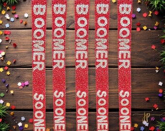 May include: Five red beaded trim with the words "Boomer Sooner" in white beads. The trim is on a wooden background with confetti.