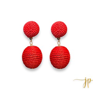 May include: A pair of red, woven ball earrings. Each earring features a small, round top and a larger, dangling sphere, connected by a gold-colored chain. The earrings are set against a clean white background.