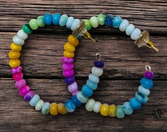 May include: Two colorful beaded hoop earrings. The earrings are made of small, round beads in a rainbow of colors, including pink, yellow, blue, and green. The earrings are displayed on a dark wooden surface.