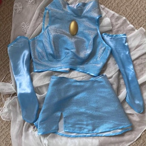 May include: A light blue costume set with a metallic sheen, including a top with a gold oval embellishment, a skirt with a side slit, and matching arm sleeves. The outfit is displayed on a white and sheer fabric.
