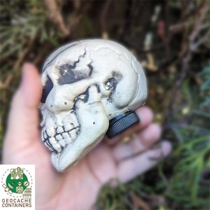 Puede incluir: Un contenedor de geocache con forma de cráneo blanco con una tapa de rosca negra. El contenedor está hecho de plástico y tiene un diseño de cráneo realista.