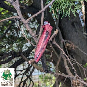 May include: A red and black geocache container shaped like a finger, placed on a tree branch. The container has a realistic appearance and is designed to blend in with its surroundings. The logo for Geocache Containers is visible in the lower left corner.