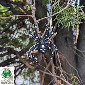May include: A black and white spider with white dots on its body is perched on a branch of a tree. The spider is made of plastic and is a toy.