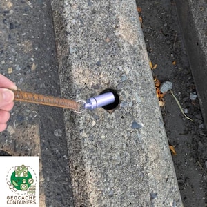 May include: A small, silver, cylindrical container is hidden in a hole in a concrete surface. A metal rod is used to retrieve the container.