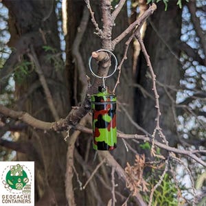Puede incluir: Un pequeño contenedor de metal de camuflaje verde, rojo y negro con un llavero plateado. El contenedor está colgado de una rama de árbol.
