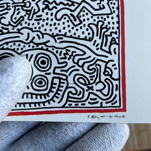 K&ouml;nnte beinhalten: Schwarz-wei&szlig;es abstraktes Kunst-Druck mit rotem Rand. Der Druck zeigt eine Vielzahl von Formen und Linien, darunter ein Gesicht mit zwei gro&szlig;en Augen. Der Druck ist unten rechts mit "K. Haring, May 7-84 K&ouml;ln" signiert.