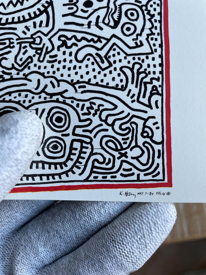 K&ouml;nnte beinhalten: Schwarz-wei&szlig;es abstraktes Kunst-Druck mit rotem Rand. Der Druck zeigt eine Vielzahl von Formen und Linien, darunter ein Gesicht mit zwei gro&szlig;en Augen. Der Druck ist unten rechts mit "K. Haring, May 7-84 K&ouml;ln" signiert.