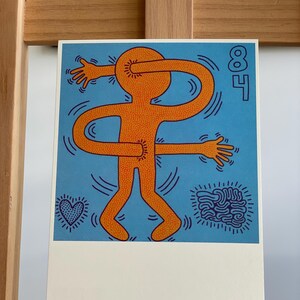 K&ouml;nnte beinhalten: Eine leuchtend orangefarbene Figur mit blauer Umrandung steht auf einem blauen Hintergrund mit einem Herz- und einem Wirbelmuster. Der Arm der Figur ist um ihren Kopf gewickelt. Die Zahl 84 befindet sich in der oberen rechten Ecke.